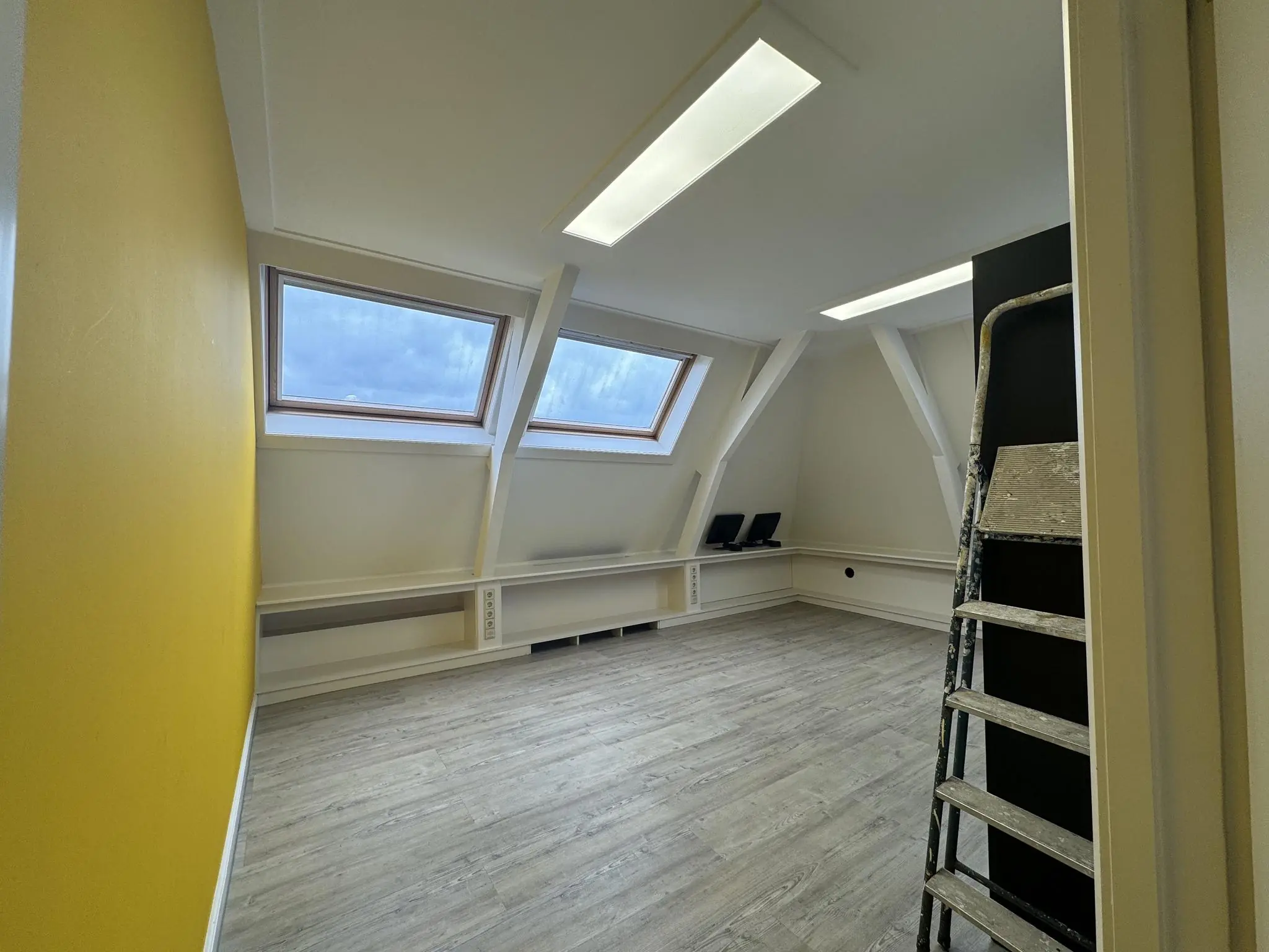 Zolderruimte met schuin dak, twee dakramen, een gele muur en een ladder tegen de wand.
